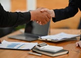 Two people shaking hands over a desk littered with notes and a calculator