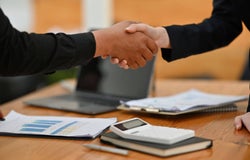 Two people shaking hands over a desk littered with notes and a calculator