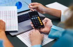 A person uses a calculator in front of a laptop in an office