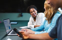 A smiling woman talks to her colleague in front of a laptop