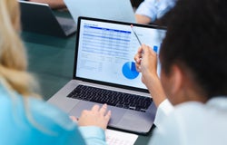 A woman points at a spreadsheet displayed on an office laptop