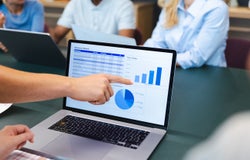 A persons hand points to a spreadsheet on a laptop screen