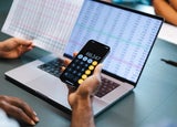 A person holds up a calculator in front of a laptop displaying a spreadsheet