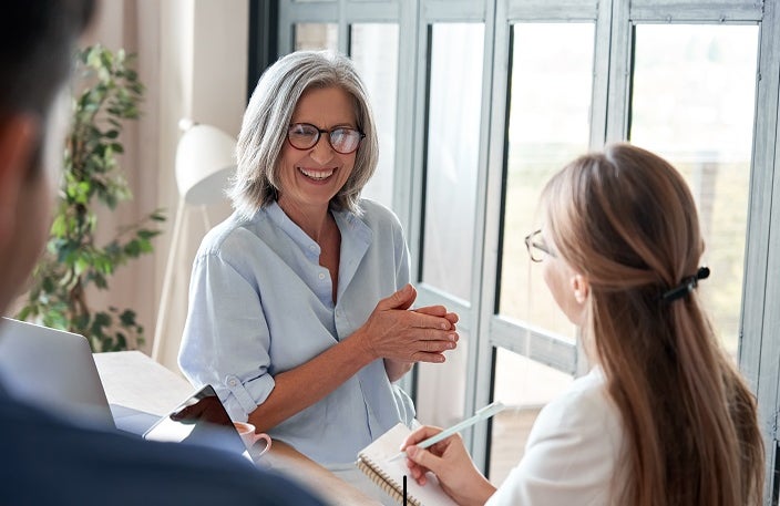 Happy mature old female mentor coach laughing training young interns at group office meeting professional workshop. Cheerful middle aged teacher professor talking to students at university seminar.