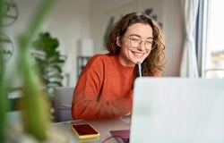 Happy young woman using laptop sitting at desk writing notes while watching webinar, studying online, looking at pc screen learning web classes or having virtual call meeting remote working from home.