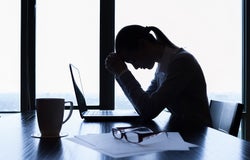 Stressed business woman sits in front of a laptop at an office desk with her head in her hands