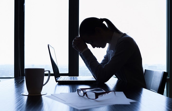 Stressed business woman sits in front of a laptop at an office desk with her head in her hands