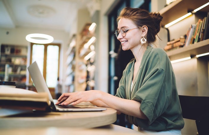 Side view of smiling adult modern female in casual outfit and with glasses sitting at table with books typing laptop against blurred shelves in modern library