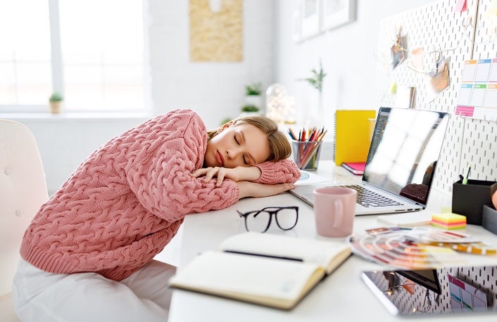 Exhausted young woman lying on table and sleeping while working on remote project in cozy home office