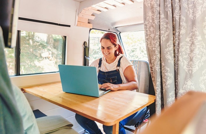 Caucasian young woman working on her laptop in her handmade camper van while traveling. Van road trip holiday and outdoor summer adventure. Nomad remote work lifestyle concept