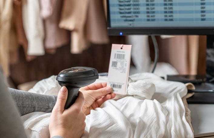 A warehouse woman employee accepts clothes using barcode scanner reading a bar code from price tag of female blouse and adds to the computer base.