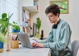 Young male college student sitting at desk at home using laptop. Handsome guy 19 years old with glasses typing on keyboard. Use internet online technologies for study, leisure, communication