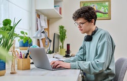Young male college student sitting at desk at home using laptop. Handsome guy 19 years old with glasses typing on keyboard. Use internet online technologies for study, leisure, communication