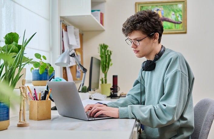 Young male college student sitting at desk at home using laptop. Handsome guy 19 years old with glasses typing on keyboard. Use internet online technologies for study, leisure, communication