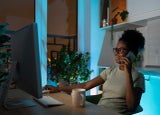 Positive woman in glasses using computer mouse, talking on smartphone, working on desktop pc late at night at home office during the night. Remote work concept