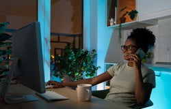 Positive woman in glasses using computer mouse, talking on smartphone, working on desktop pc late at night at home office during the night. Remote work concept