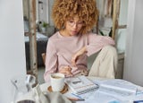 Serious curly haired woman manages household family budget calculates expenditures takes care of finances and savings sits at table with receipts dressed in domestic clothes poses at home office