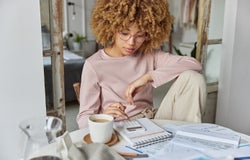 Serious curly haired woman manages household family budget calculates expenditures takes care of finances and savings sits at table with receipts dressed in domestic clothes poses at home office