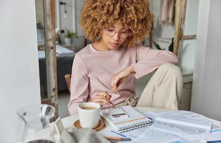 Serious curly haired woman manages household family budget calculates expenditures takes care of finances and savings sits at table with receipts dressed in domestic clothes poses at home office