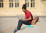 Male Student Working On Laptop Sitting On StepsOutside University Building In Oxford UK