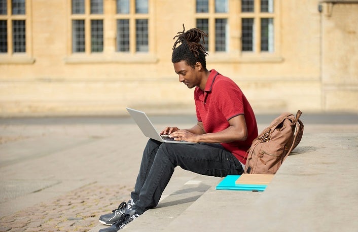 Male Student Working On Laptop Sitting On StepsOutside University Building In Oxford UK