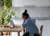 Unhappy young woman sitting at kitchen table looking at laptop having problems in job searching