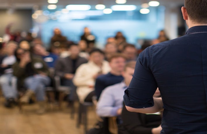 Back of a man presenting to a group of seated people in soft focus