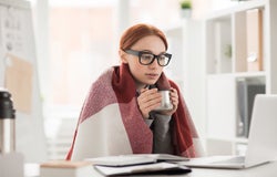 Young woman in plaid and eyeglasses drinking hot tea from mug during work in office