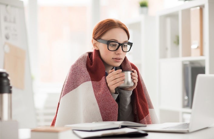 Young woman in plaid and eyeglasses drinking hot tea from mug during work in office