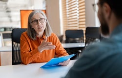 Senior business woman, employer holding resume listening young candidate at job interview. Group of business people talking, meeting, sharing ideas in modern office. Human resources concept
