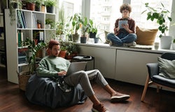 Young woman with tablet sitting on windowsill in front of male colleague with laptop networking in armchair during individual work
