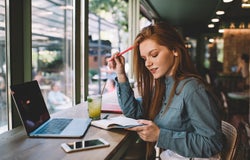 Side view of young female freelancer with pen touching hair and looking down at notebook while sitting at desk with laptop smartphone lemonade glass and worried about work progress in blurred cafe