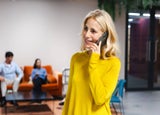 Woman standing in an office using her personal phone to make a call while colleagues work behind her