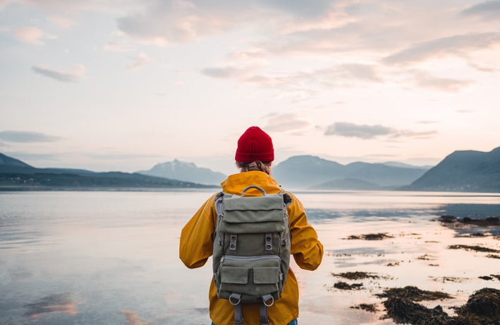 Back view of male tourist with rucksack standing on coast in front of great mountain massif while journey. Man traveler wearing yellow jacket with backpack explore scandinavia nature. Wanderlust outdoor lifestyle