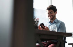 Man sitting at a desk, looking at his smartphone and smiling