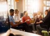 Group of employees sitting around the office chatting and laughing with mugs of coffee.