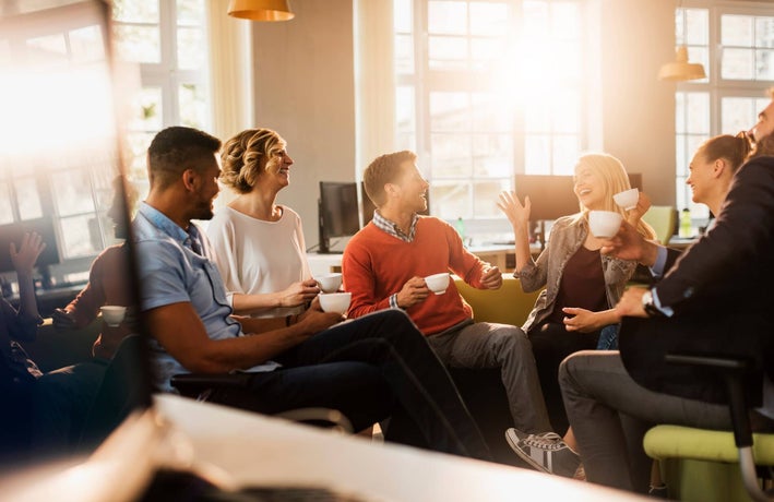 Group of employees sitting around the office chatting and laughing with mugs of coffee.