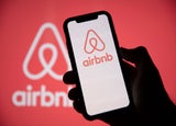 Silhouette of a hand holding up a smartphone displaying the AirBnB logo on its screen against a red backdrop.