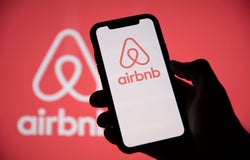 Silhouette of a hand holding up a smartphone displaying the AirBnB logo on its screen against a red backdrop.