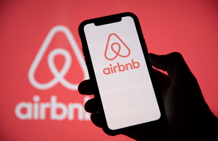 Silhouette of a hand holding up a smartphone displaying the AirBnB logo on its screen against a red backdrop.