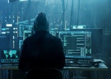 Backshot of a hooded person in a dark room with multiple computer screens.