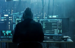 Backshot of a hooded person in a dark room with multiple computer screens.
