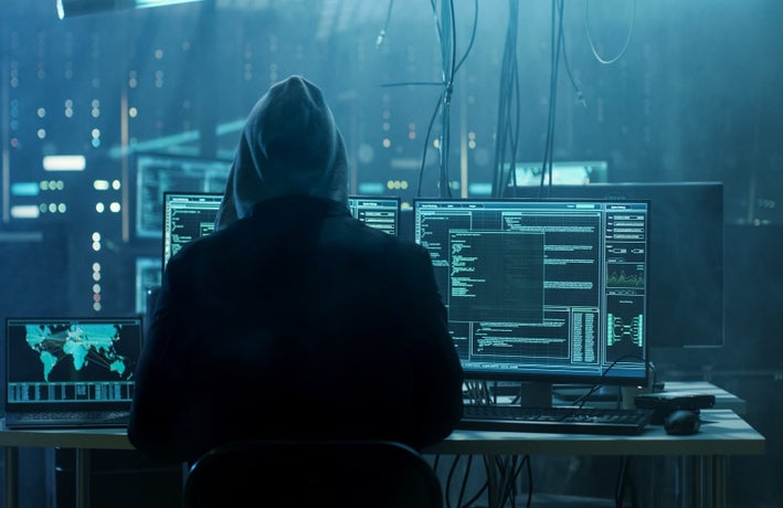 Backshot of a hooded person in a dark room with multiple computer screens.