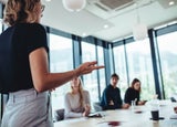 A back shot of a blonde woman in a black top and grey trousers addressing team members in a meeting room.
