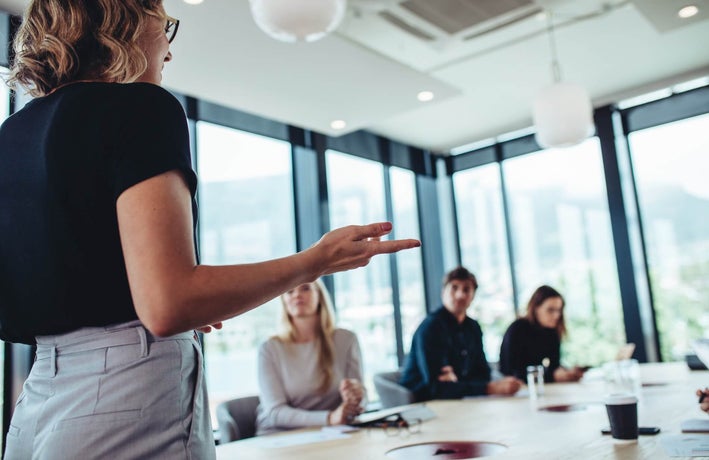 A back shot of a blonde woman in a black top and grey trousers addressing team members in a meeting room.