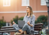 Lady meditating on an urban bench with a laptop perched between her knees.