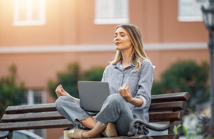 Lady meditating on an urban bench with a laptop perched between her knees.