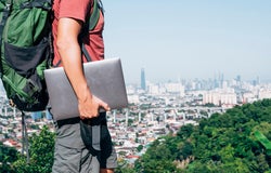 A man wearing a red shirt, cargo shorts and large green backpack walking along the cityscape with a laptop in his right arm.