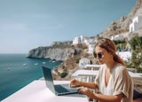 A lady with a loose ponytail and sunglasses sitting outside a cafe at her laptop with a view of the ocean.