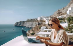 A lady with a loose ponytail and sunglasses sitting outside a cafe at her laptop with a view of the ocean.